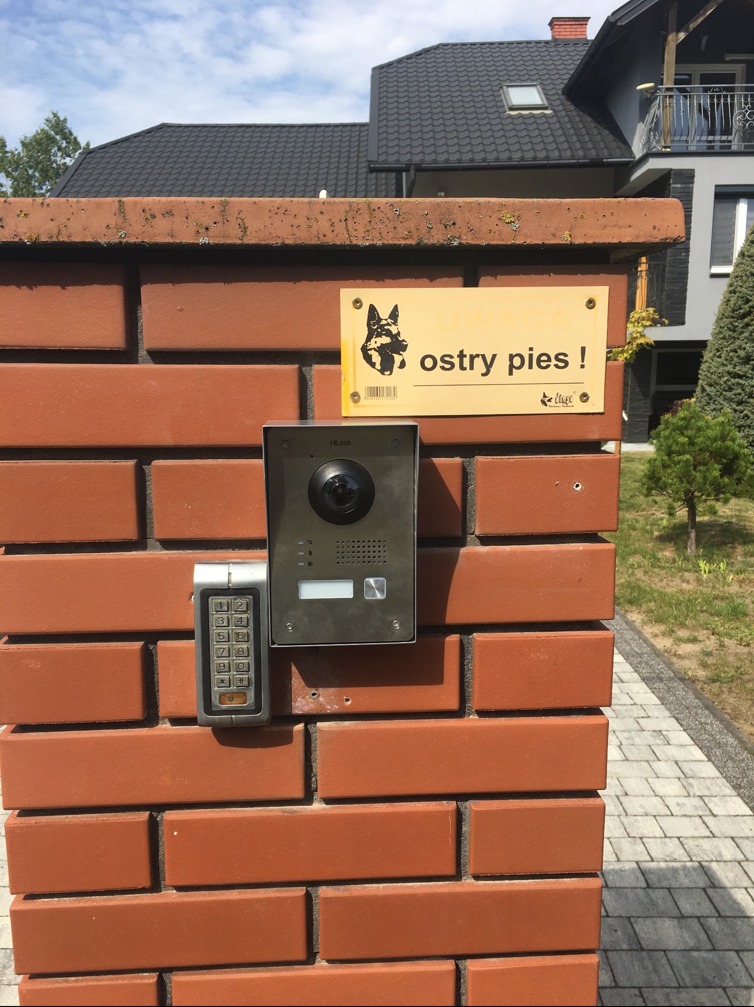 Zewnętrzny panel wideodomofonu z klawiaturą numeryczną, zamontowany na ceglanym słupku ogrodzenia obok tabliczki ostrzegającej przed psem.