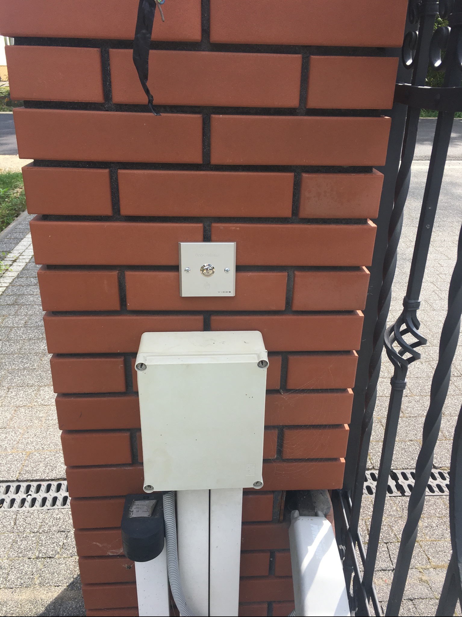 Domofon Vidos zamontowany na ceglanym słupku ogrodzenia, obok skrzynka instalacyjna i brama z metaloplastyki