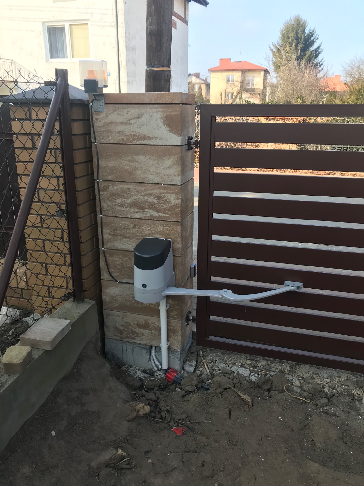 Automatyczna brama wjazdowa z brązowymi poziomymi panelami, napędem zamontowanym na murowanym słupku z kostki, widoczne przewody i elementy instalacji elektrycznej.