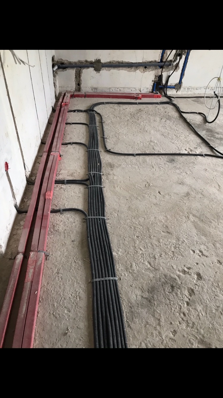 Kable elektryczne w peszlach ułożone równolegle na surowym betonowym podłożu w trakcie prac instalacyjnych, zabezpieczone metalowymi opaskami; widoczne również czerwone koryta kablowe przy ścianie.