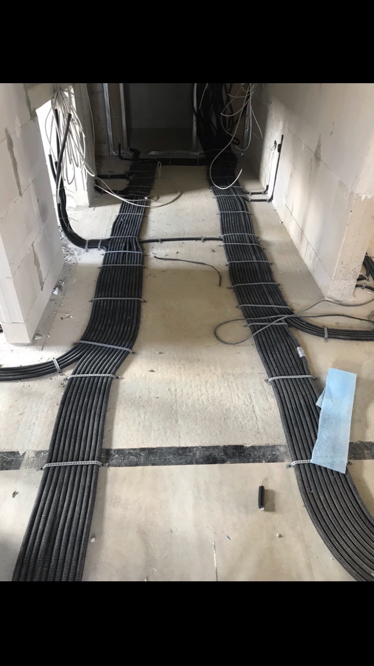 Równolegle ułożone wiązki peszli z kablami elektrycznymi, przymocowane metalowymi opaskami do surowej posadzki w trakcie instalacji w nowym budynku.