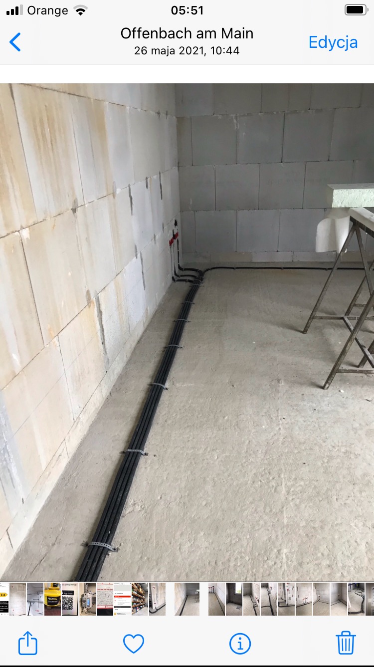 Surowy stan: cztery czarne kable elektryczne przymocowane do ściany w nowo budowanym pomieszczeniu z bloków betonowych, widoczne czerwone rurki instalacyjne i metalowa drabina w tle.