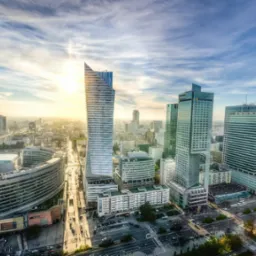 Panorama Warszawy z widokiem na drapacze chmur w promieniach zachodzącego słońca, widoczny ruch uliczny i charakterystyczna architektura miejska.