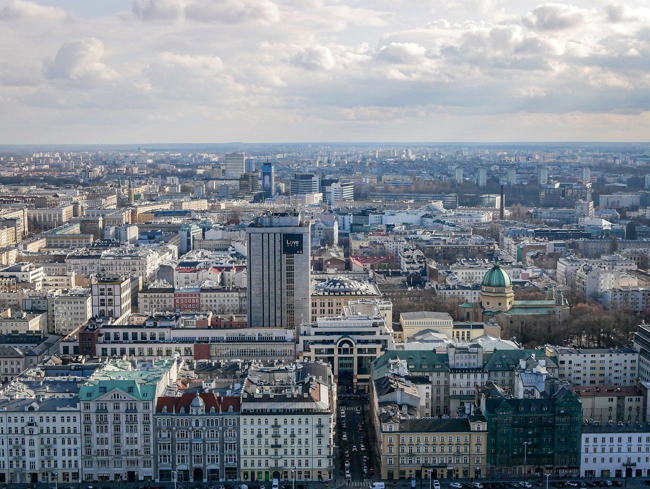 Panorama Warszawy z lotu ptaka, widoczne budynki mieszkalne i usługowe, kościół z zieloną kopułą, pochmurne niebo.