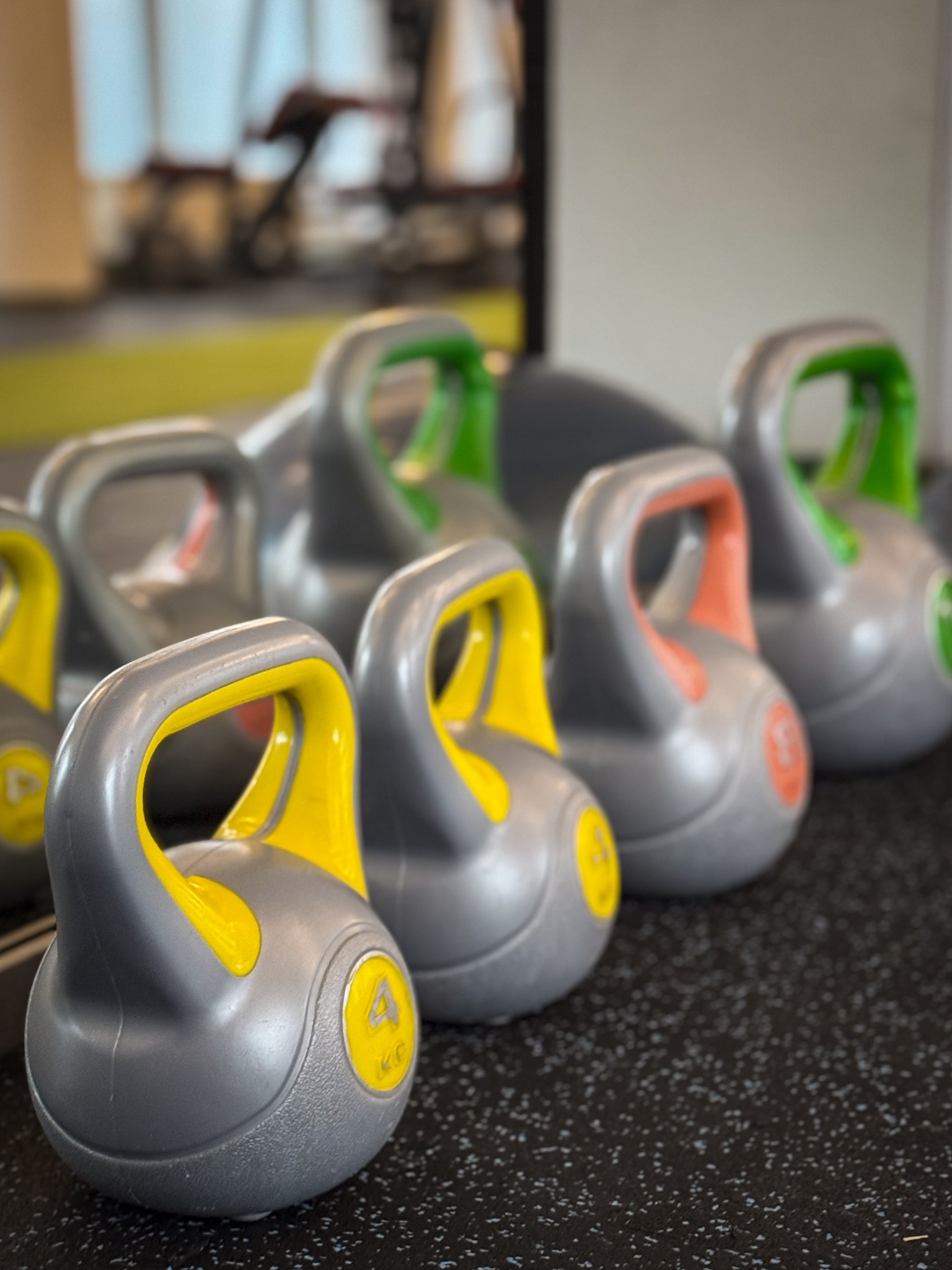 Rząd szarych kettlebell z kolorowymi uchwytami (żółtym, pomarańczowym, zielonym) ułożonych na czarnej podłodze w siłowni. Widoczny napis 4 KG na pierwszym planie.