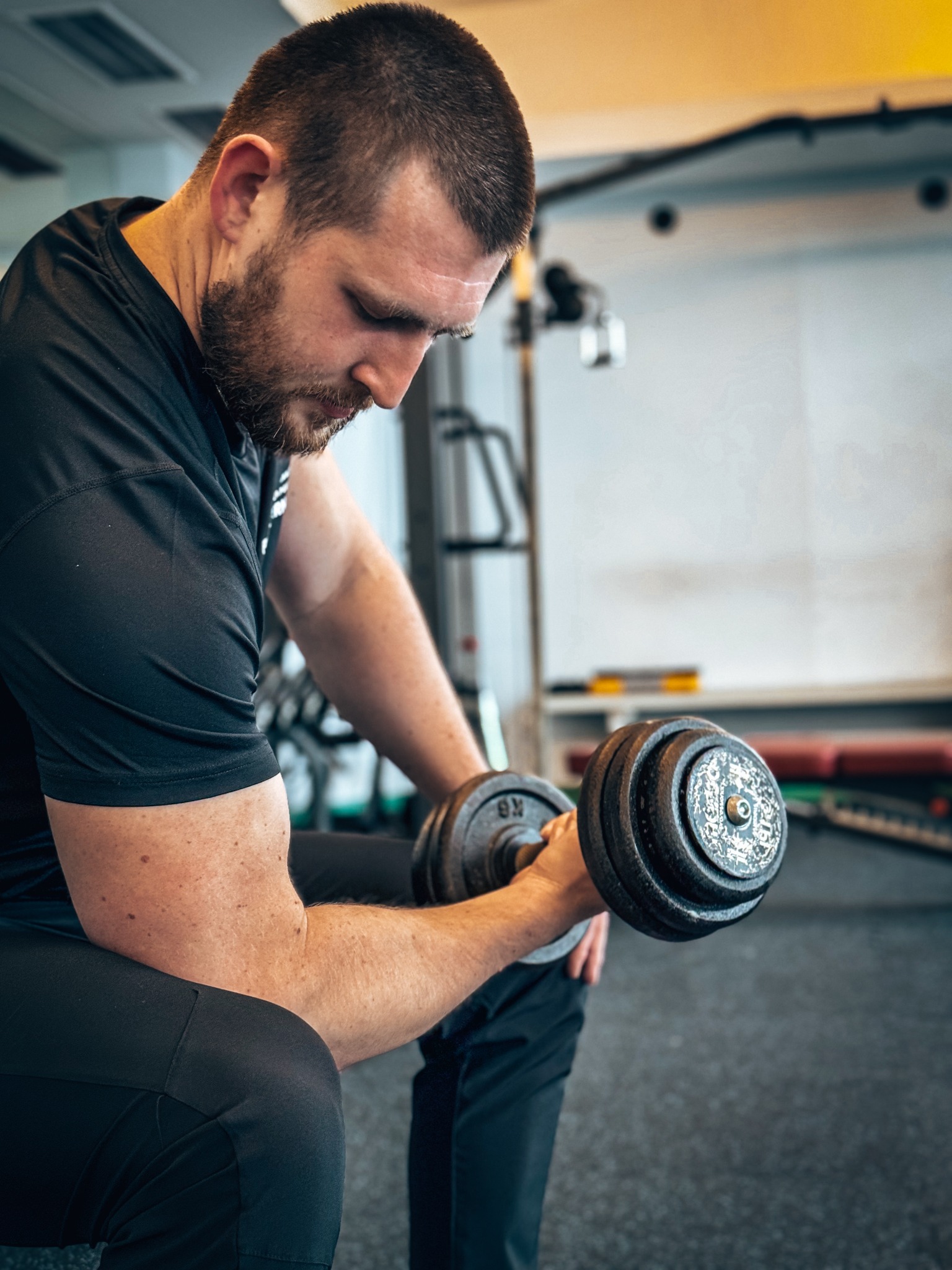 Mężczyzna w czarnym stroju sportowym, siedząc na ławce, wykonuje ćwiczenie bicepsa z hantlą w siłowni.