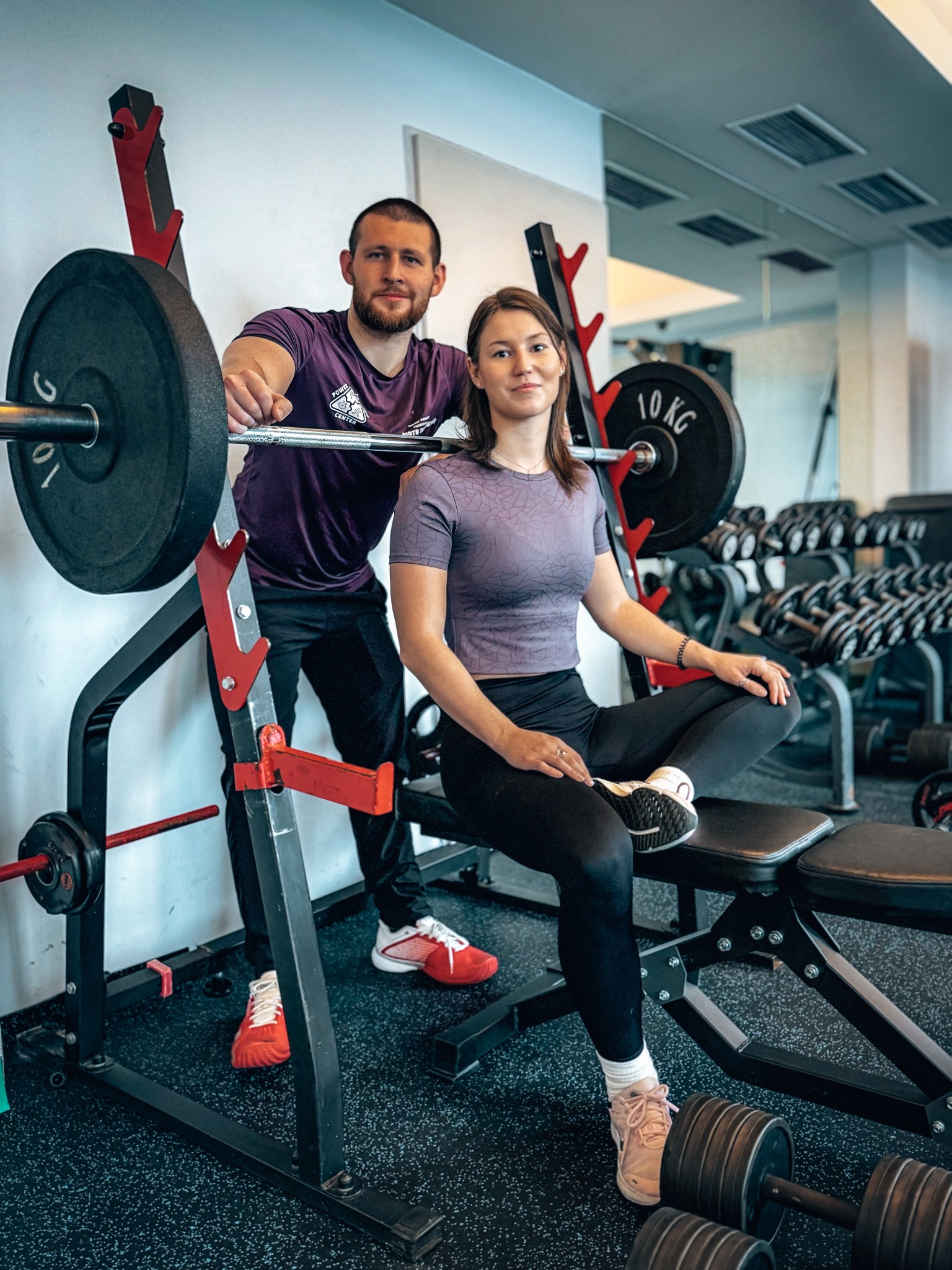 Kobieta siedzi na ławce do ćwiczeń, a mężczyzna w fioletowej koszulce stoi obok z sztangą na stojaku w siłowni.
