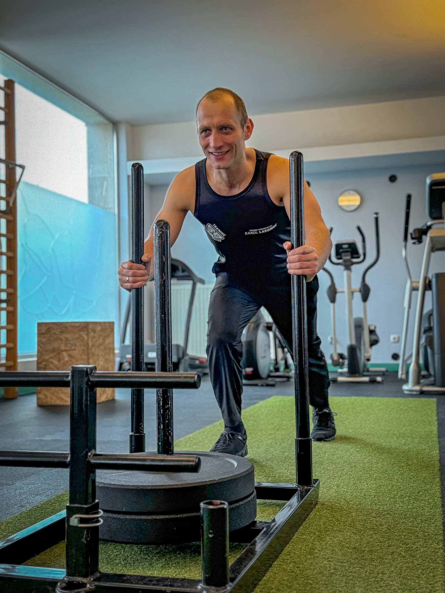 Uśmiechnięty mężczyzna w czarnym stroju sportowym pcha obciążony wózek treningowy na zielonej sztucznej trawie w siłowni, widoczne inne urządzenia do ćwiczeń.