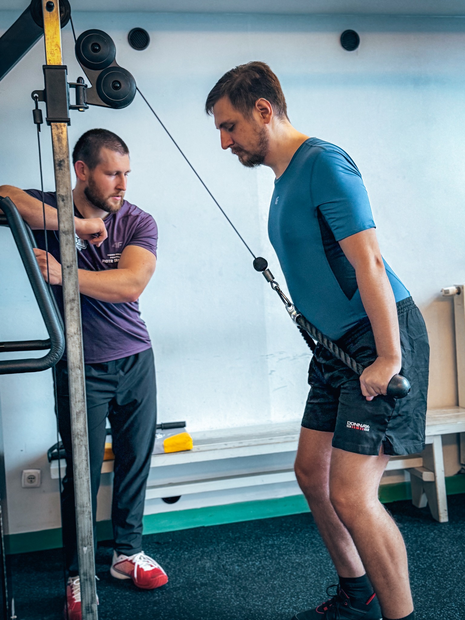Mężczyzna w niebieskiej koszulce ćwiczy na maszynie pod nadzorem drugiego mężczyzny w fioletowej koszulce, który opiera się o konstrukcję maszyny.