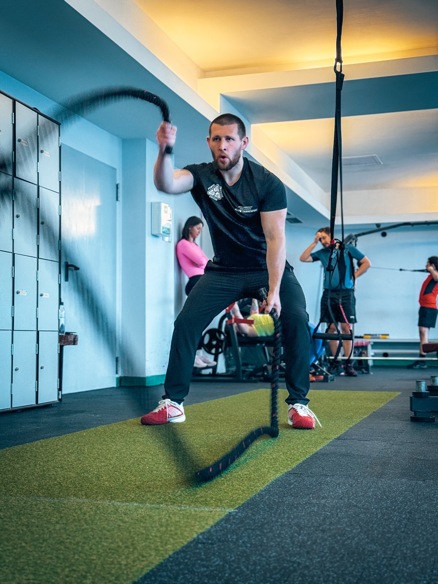 Mężczyzna w czarnej koszulce z logiem, ćwiczący z linami w siłowni, w tle szafki i inne osoby.