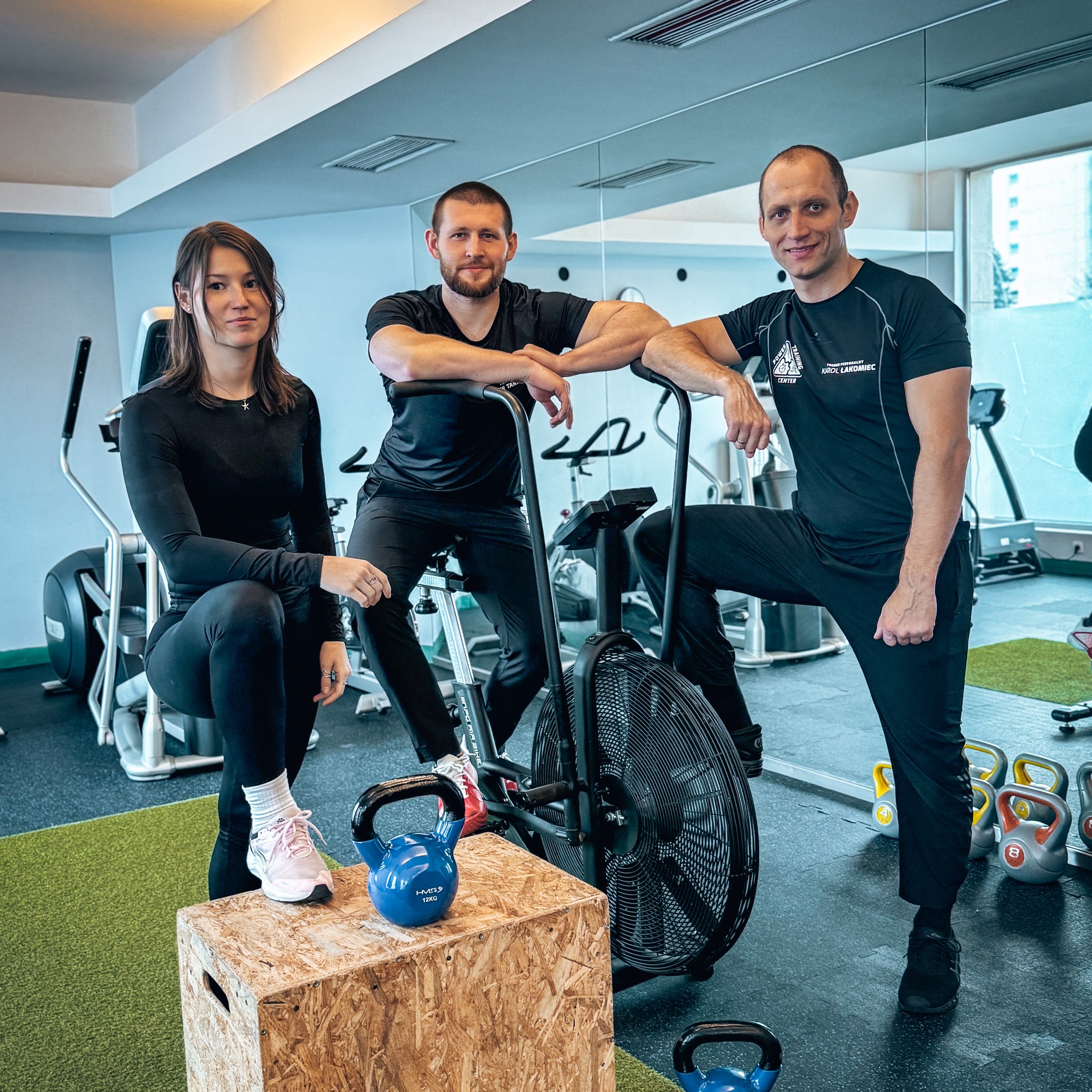 Trzy osoby w czarnych strojach sportowych pozują w siłowni obok roweru treningowego, drewnianego podestu i kettlebelli; jeden z mężczyzn ma koszulkę z logo.