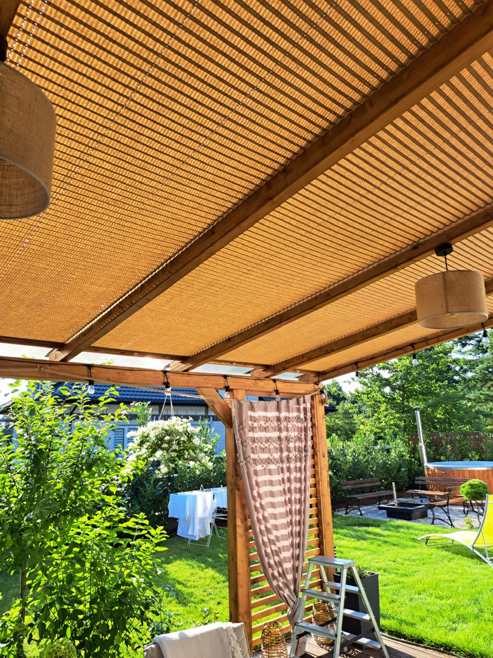 Zadaszenie tarasu z beżową tkaniną w paski, drewniana konstrukcja, widok na ogród z jacuzzi i suszącym się praniem.