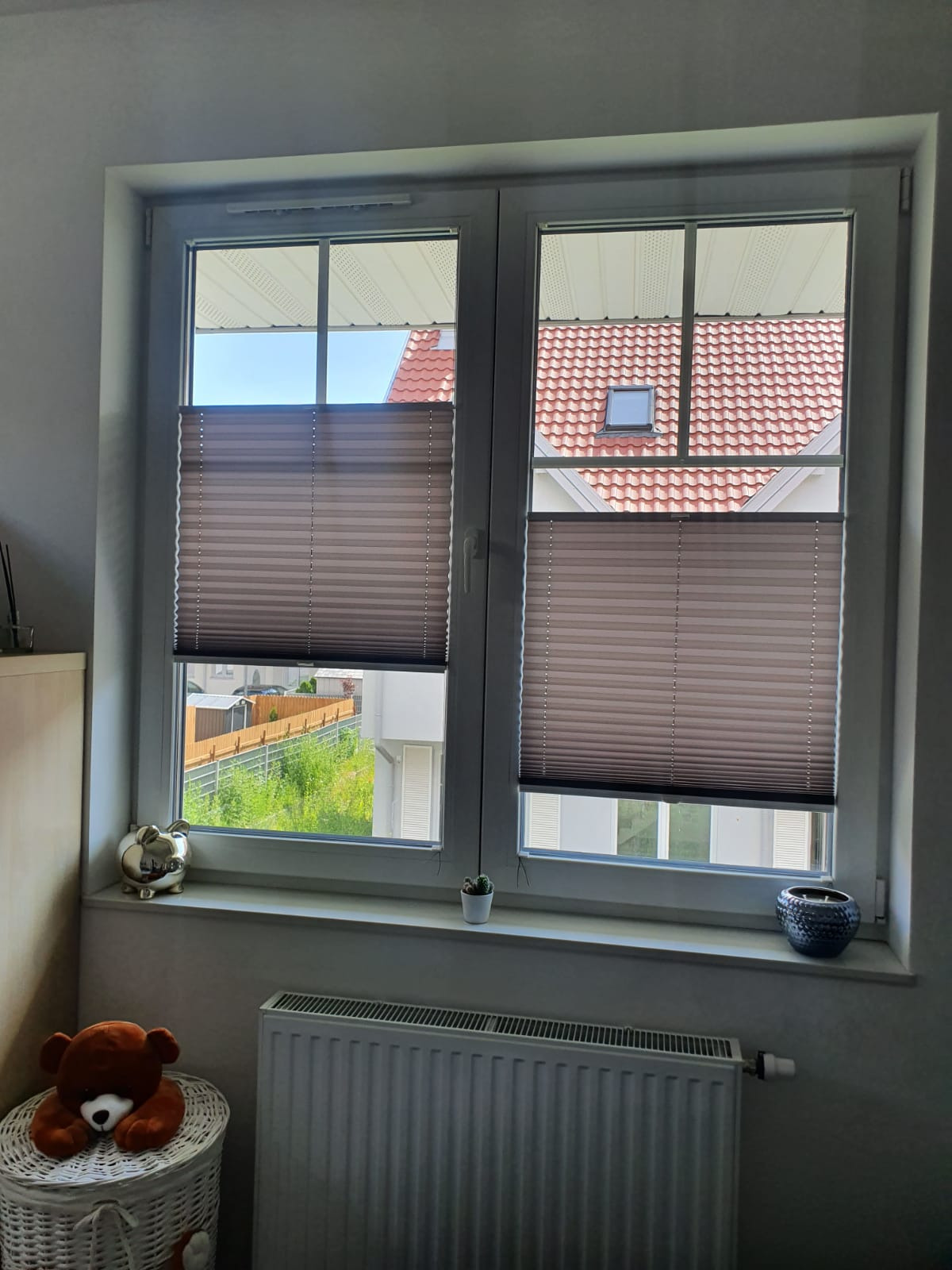 Brązowe, częściowo zasłonięte okno z plisami, widok na dach domu i zieleń, na parapecie srebrna świnka skarbonka i mały kaktus, w pokoju pluszowy miś i biały grzejnik.