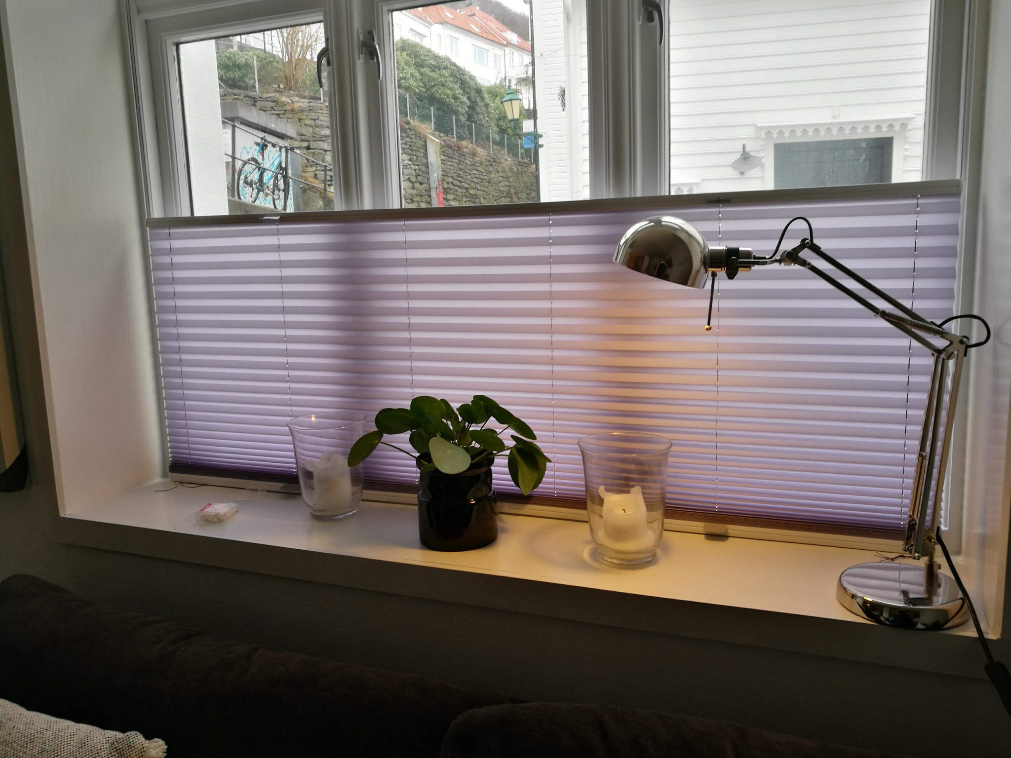 Jasnofioletowe, poziome osłony okienne w formie harmonijki, widoczne z wnętrza, z lampką biurkową i dekoracjami na parapecie, z widokiem na zabudowę mieszkalną za oknem.