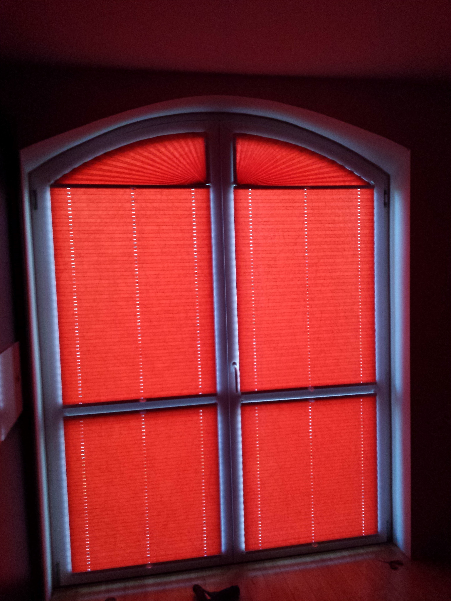 Czerwone, plisowane osłony okienne w łukowym oknie, widoczne z dołu, z delikatnym odbiciem światła na dolnej krawędzi.