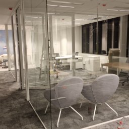 GRUPA DB - Wnętrze biura w Warszawie z przeszklonymi ścianami działowymi, widoczne biurka, krzesła, wykładzina dywanowa oraz torba robocza przy ścianie, sugerujące prace wykończeniowe.