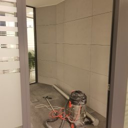 GRUPA DB - Wnętrze biura w trakcie sprzątania: odkurzacz przemysłowy z czerwonymi elementami i wężem na szarym dywanie, widok przez otwarte drzwi z matowym szkłem.