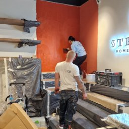 GRUPA DB - Pracownicy nakładają dekoracyjną pomarańczową farbę strukturalną na ścianę w biurze, widoczne narzędzia, częściowo zabezpieczone elementy wyposażenia i oświetlenie sufitowe.