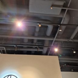 GRUPA DB - Szare rury wentylacyjne i oświetlenie sufitowe w biurze, widoczny fragment podświetlanego logo firmy na ścianie.