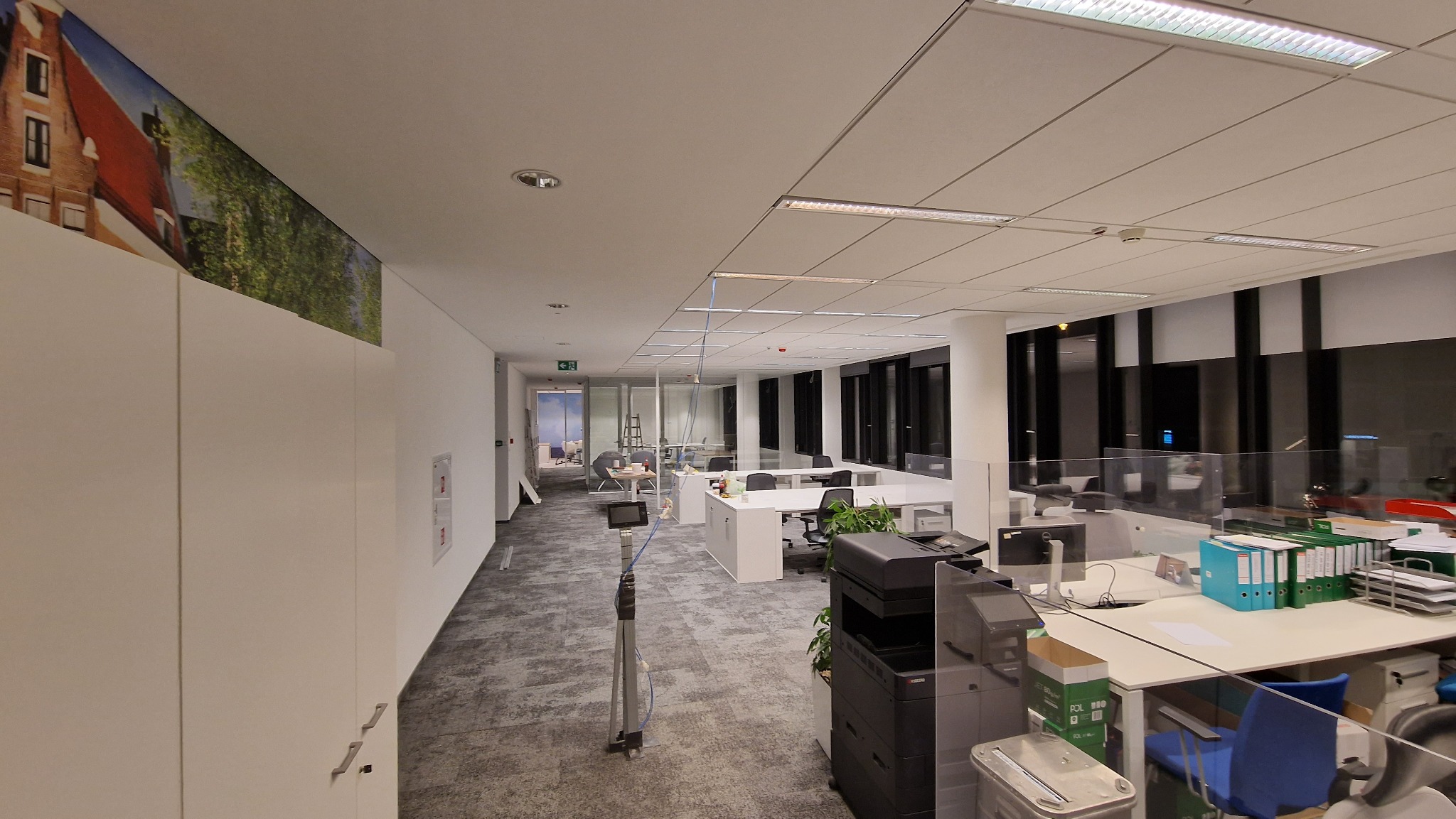 Przestronne biuro z białymi biurkami, krzesłami i roślinami doniczkowymi, oddzielone szklanymi przegrodami, z widocznym fragmentem fototapety przedstawiającej budynki w tle