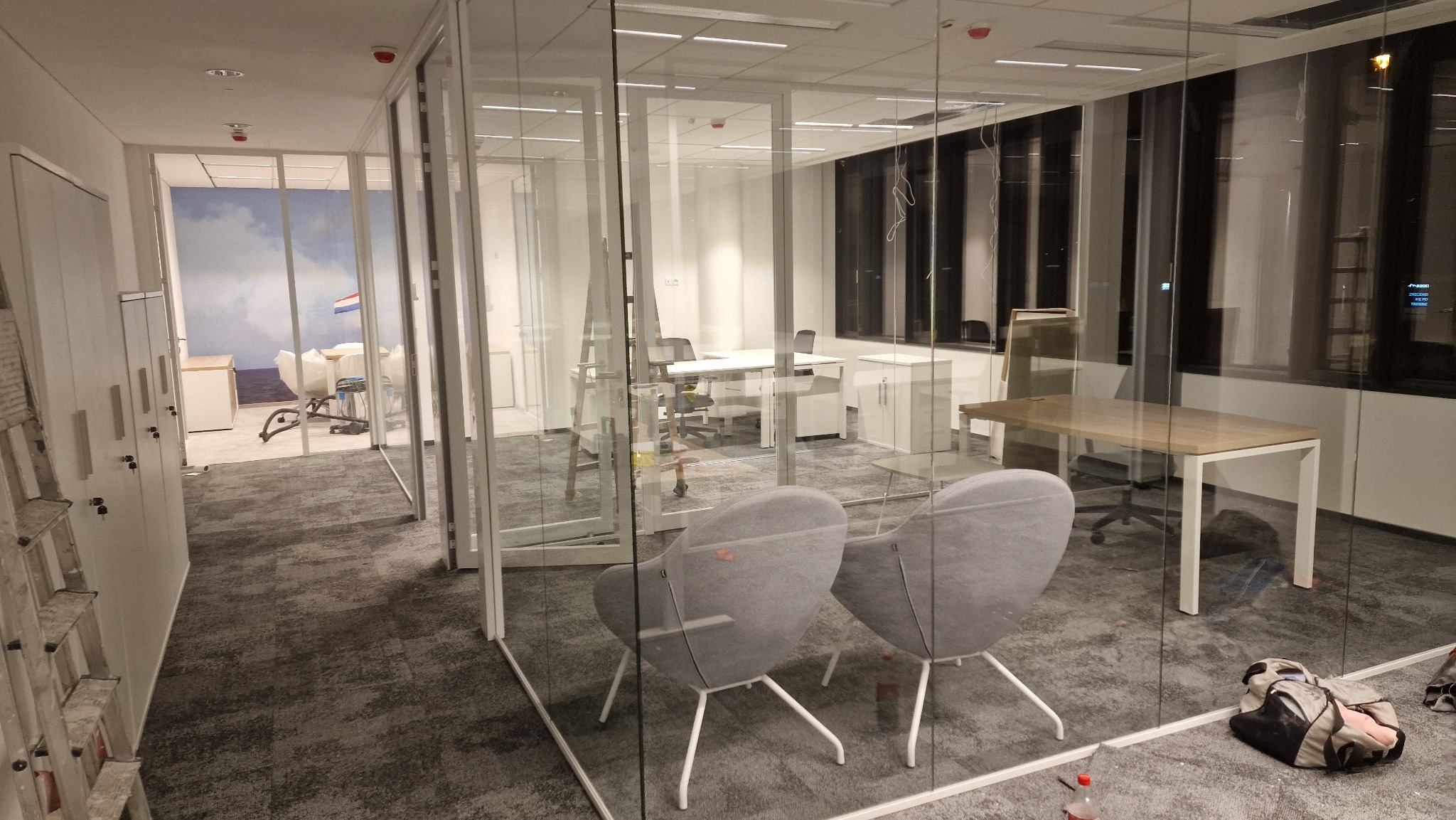 Wnętrze biura w Warszawie z przeszklonymi ścianami działowymi, widoczne biurka, krzesła, wykładzina dywanowa oraz torba robocza przy ścianie, sugerujące prace wykończeniowe.