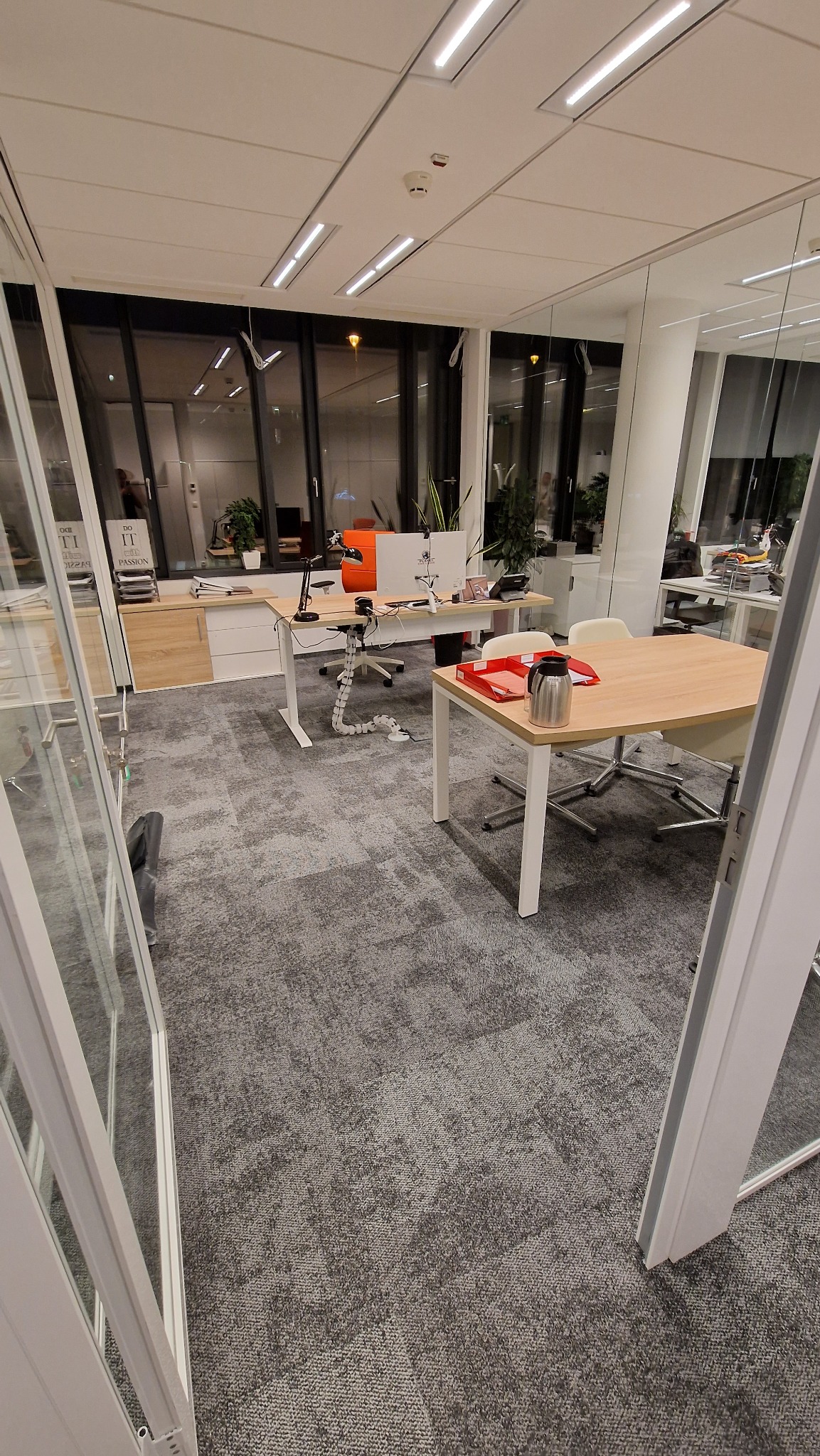 Jasne biuro z widocznymi biurkami, krzesłami, wykładziną dywanową i roślinami doniczkowymi, widziane przez otwarte szklane drzwi.