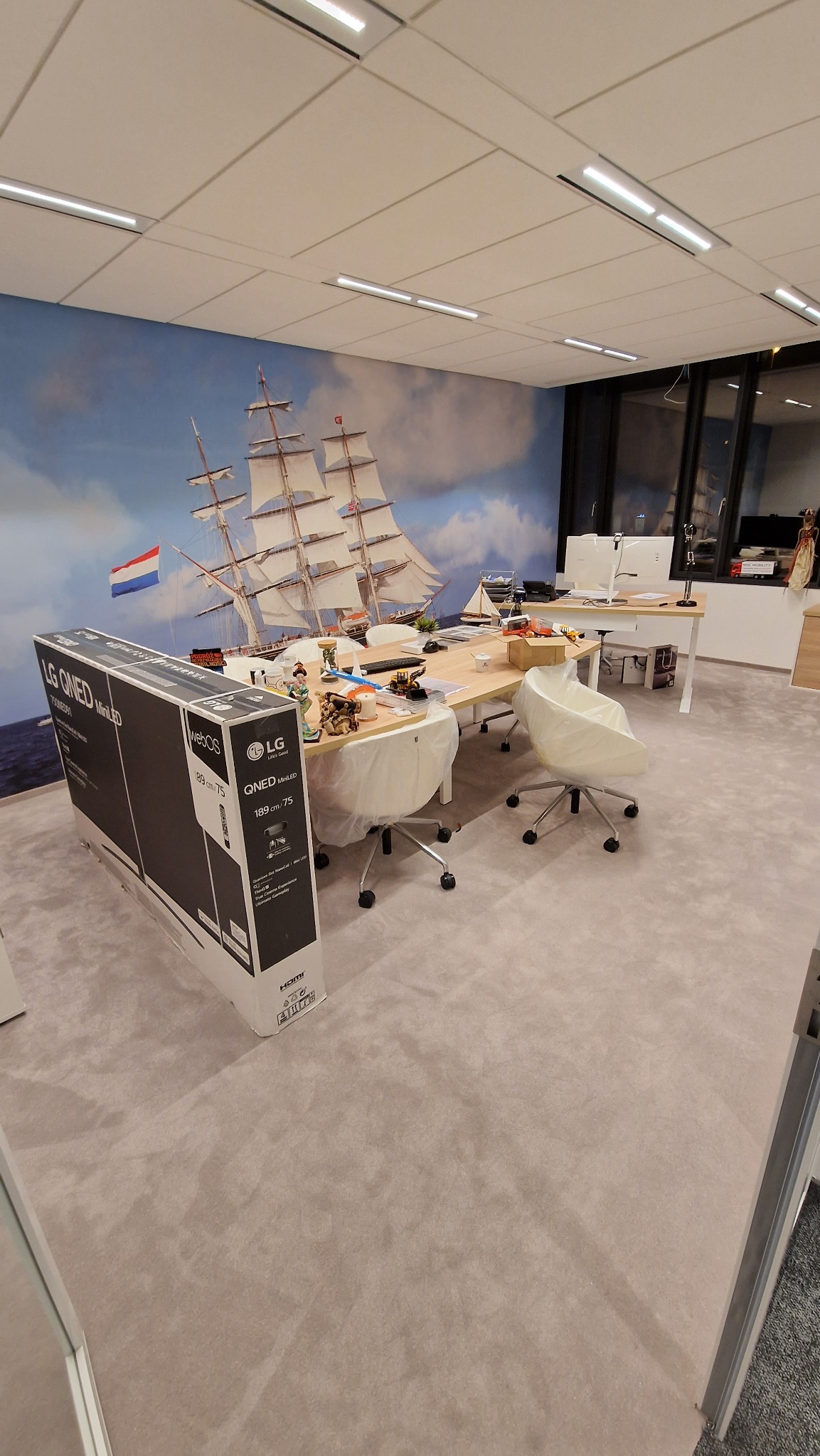 Biuro po remoncie z fototapetą przedstawiającą żaglowiec, karton nowego telewizora LG QNED MiniLED 75 cali stoi na podłodze, biurka częściowo zakryte folią ochronną, białe krzesła na kółkach.