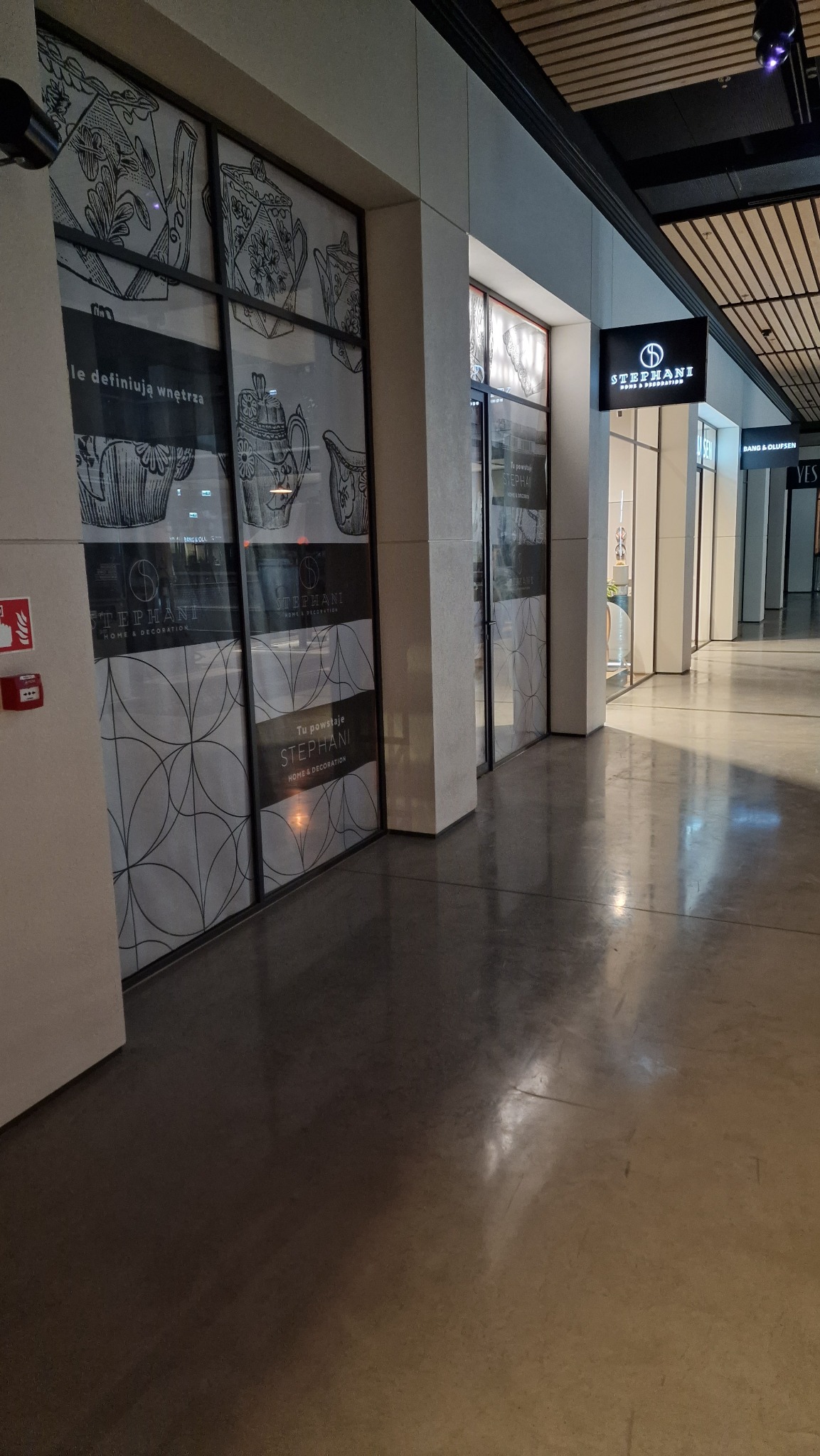 Wnętrze galerii handlowej z widokiem na witrynę sklepu z dekoracjami wnętrz, oklejona grafiką z motywem dzbanków i czajników, betonowa podłoga.