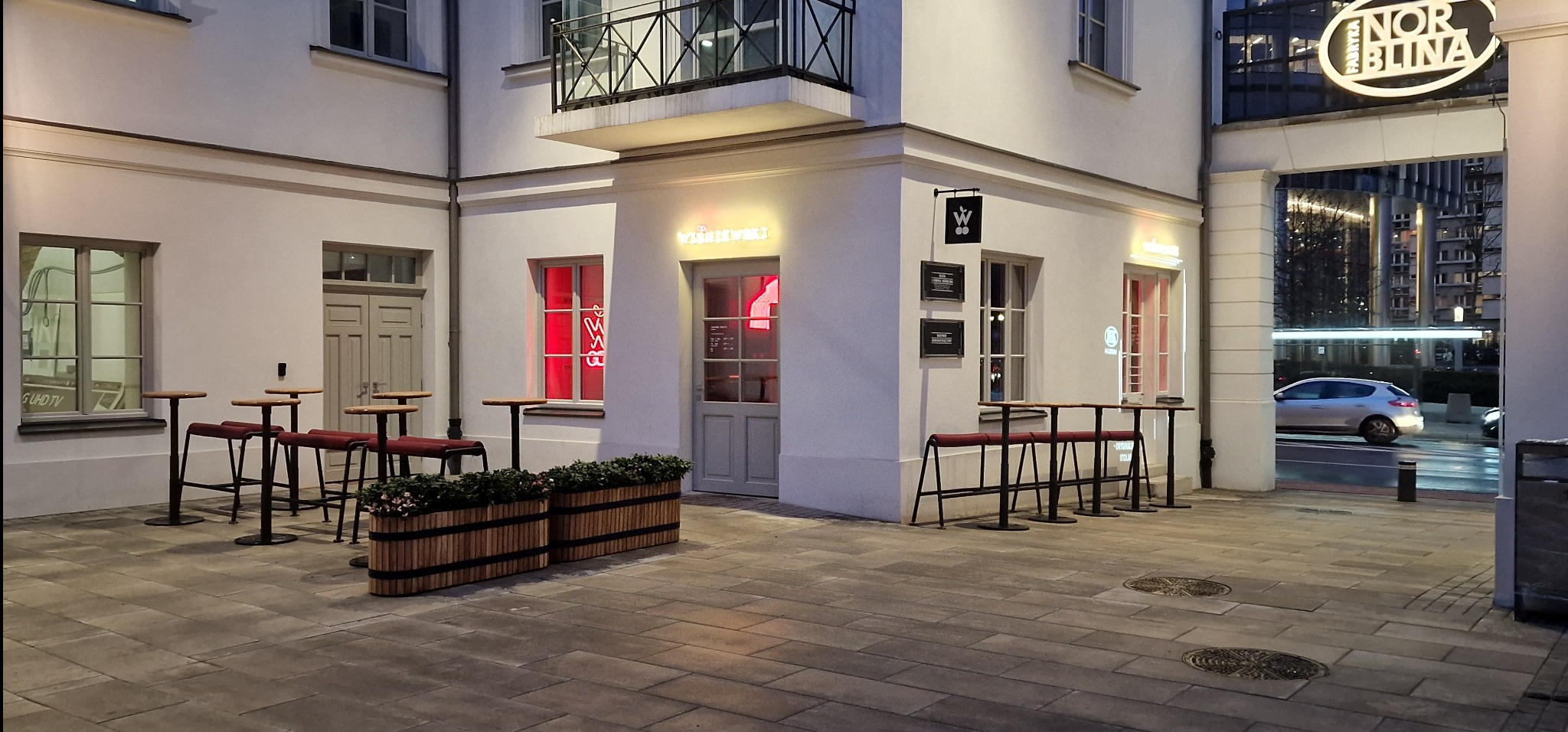 Elewacja budynku z neonowym napisem i logo, stoliki barowe na zewnątrz, widok na ulicę i budynki w tle.