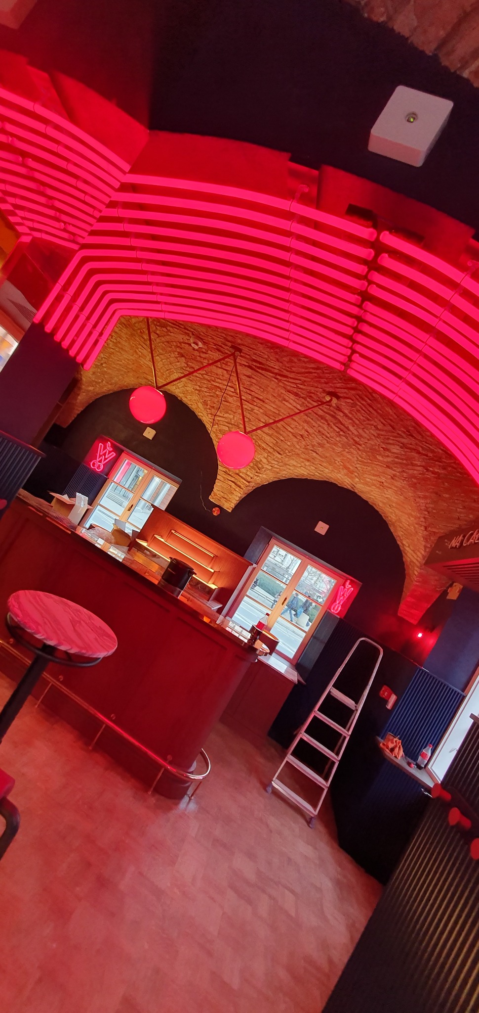 Wnętrze lokalu z ceglanym sklepieniem oświetlonym neonowym, czerwonym światłem, widoczny bar, krzesło barowe, drabina i dekoracje neonowe