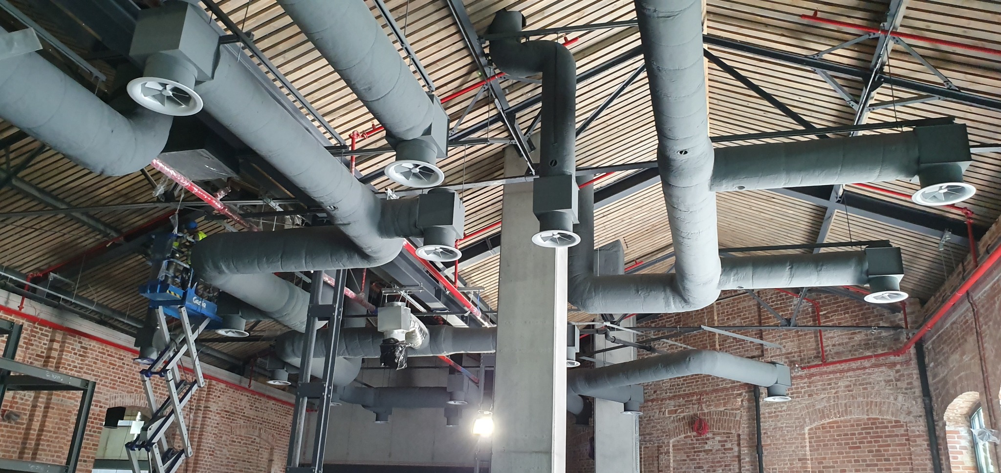 Malowanie rur wentylacyjnych na szaro w biurze z odsłoniętą cegłą, widoczny podnośnik koszowy.