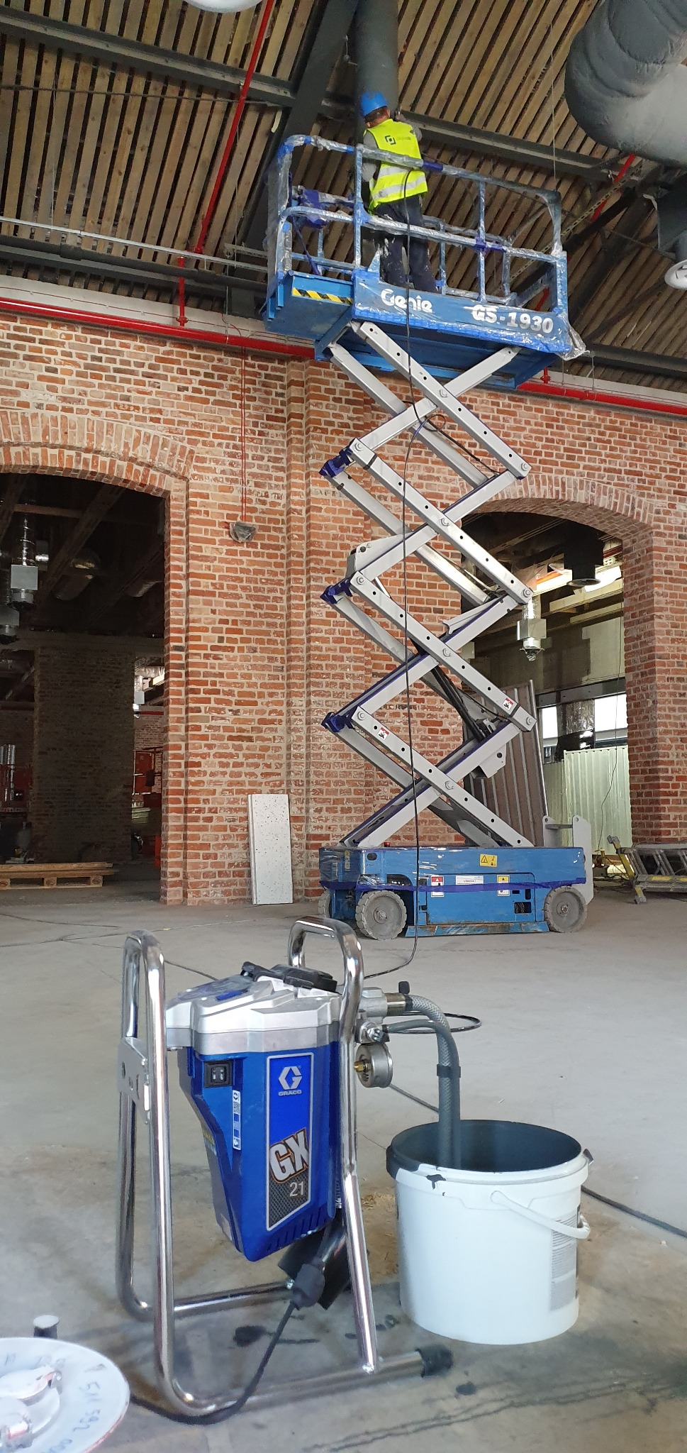 Pracownik na podnośniku nożycowym maluje rury pod sufitem w pomieszczeniu z ceglanymi ścianami, w tle widoczny agregat malarski Graco GX 21 i białe wiadro.