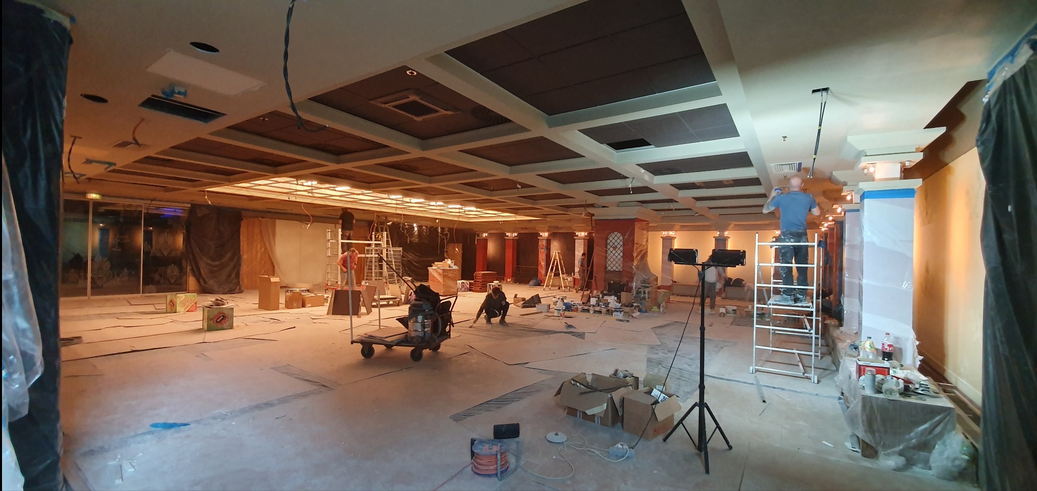 Przestronne wnętrze biurowe w trakcie prac wykończeniowych: malowanie sufitu kasetonowego, zabezpieczanie kolumn folią, rozstawione drabiny i sprzęt oświetleniowy.
