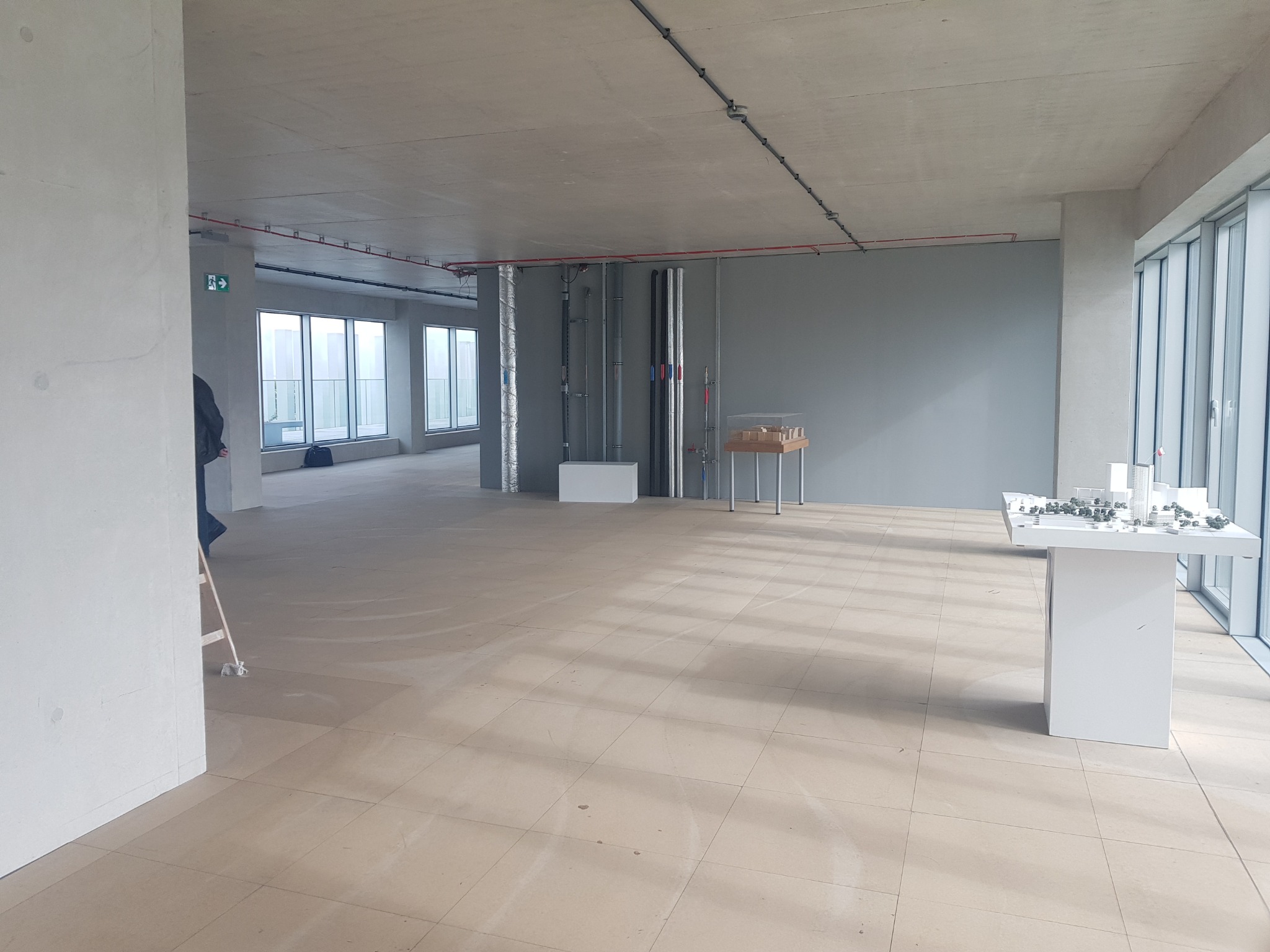 Surowe wnętrze biurowe w stanie deweloperskim z widocznymi instalacjami, modelami architektonicznymi na stolikach i fragmentem drabiny malarskiej przy ścianie.