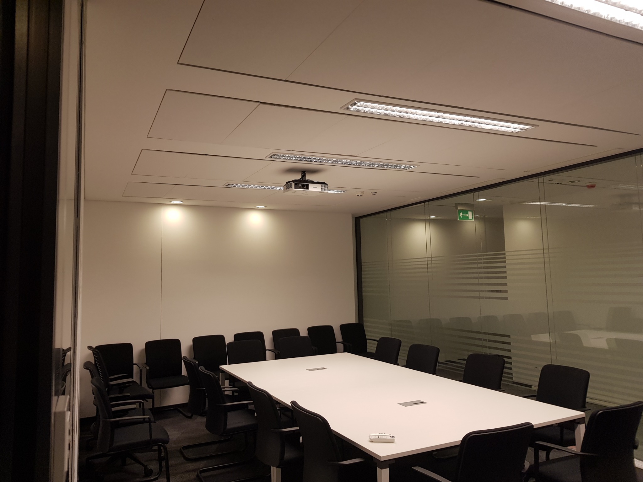 Sala konferencyjna z białym stołem, czarnymi krzesłami i projektorem zamontowanym na suficie. Ściana ze szkła z poziomymi paskami.