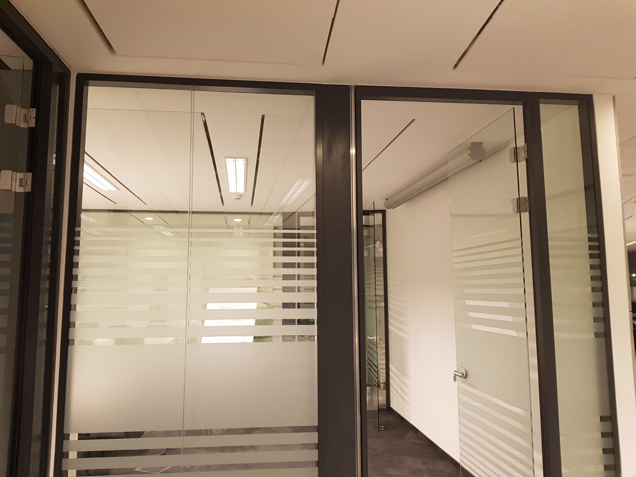 Szklane drzwi biurowe z poziomymi, matowymi pasami, widoczne z perspektywy korytarza, z elementami oświetlenia sufitowego w tle.
