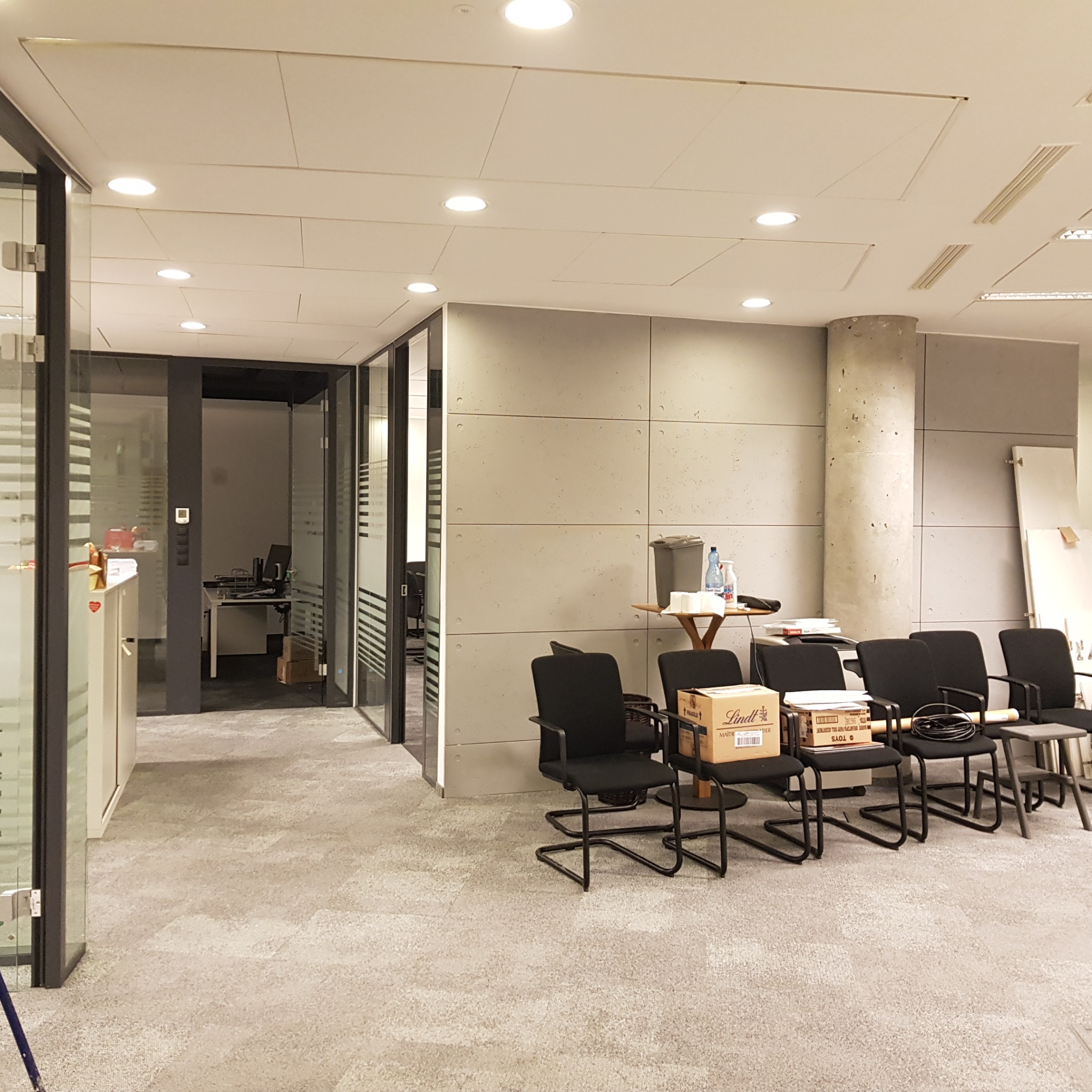 Przygotowane do użytku, ale jeszcze puste biuro z szarymi ścianami, krzesłami z pudełkami, przeszklonymi ścianami działowymi i wykładziną dywanową w odcieniach szarości.