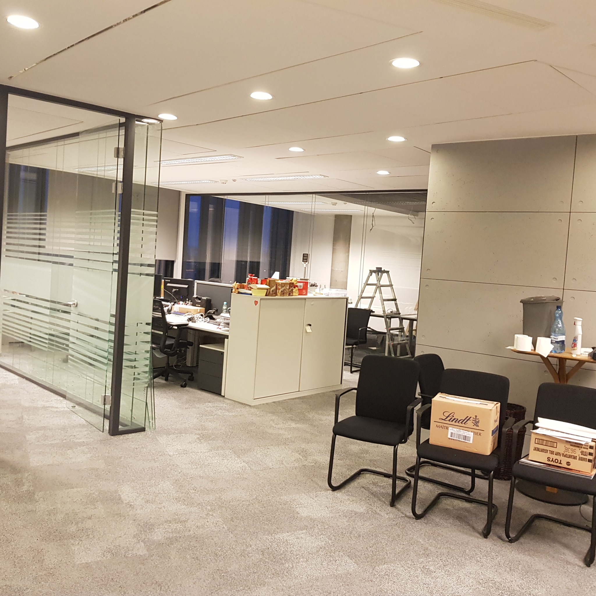 Biuro w trakcie odświeżania: na krzesłach kartony, w tle drabina malarska, widoczne ślady prac na ścianie i suficie, przeszklone ścianki działowe.