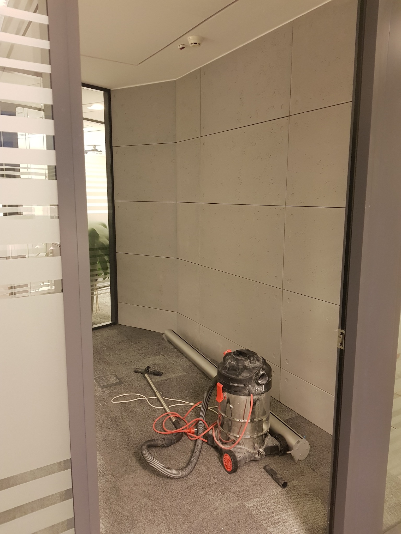 Wnętrze biura w trakcie sprzątania: odkurzacz przemysłowy z czerwonymi elementami i wężem na szarym dywanie, widok przez otwarte drzwi z matowym szkłem.