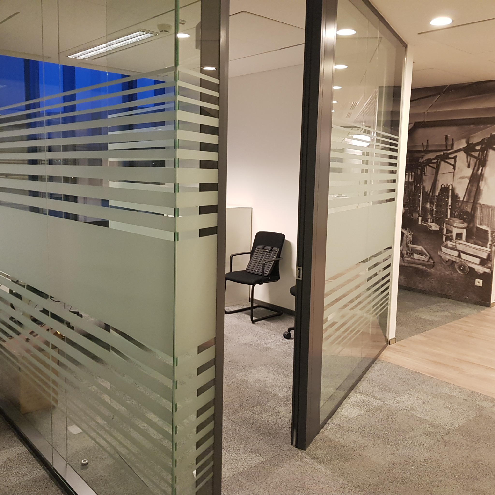 Nowoczesne biuro z przeszklonymi ścianami z poziomymi pasami, widok na małe pomieszczenie z czarnym krzesłem i klawiaturą na siedzisku, w tle fototapeta z motywami przemysłowymi.