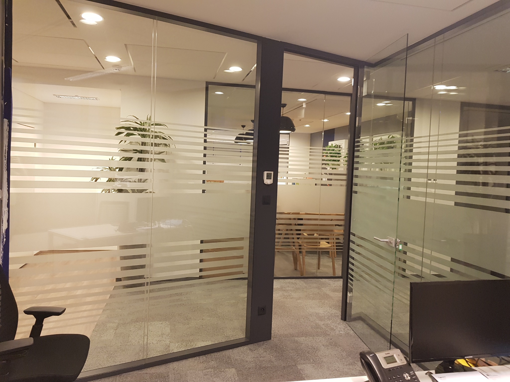 Przeszklona ściana działowa w biurze z poziomymi pasami folii, widok na salę konferencyjną z jasnymi krzesłami i rośliną doniczkową.