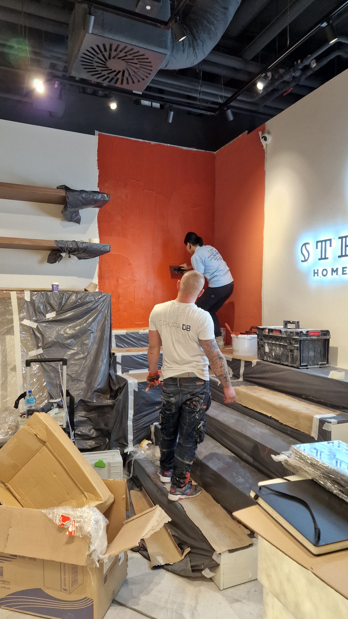 Pracownicy nakładają dekoracyjną pomarańczową farbę strukturalną na ścianę w biurze, widoczne narzędzia, częściowo zabezpieczone elementy wyposażenia i oświetlenie sufitowe.