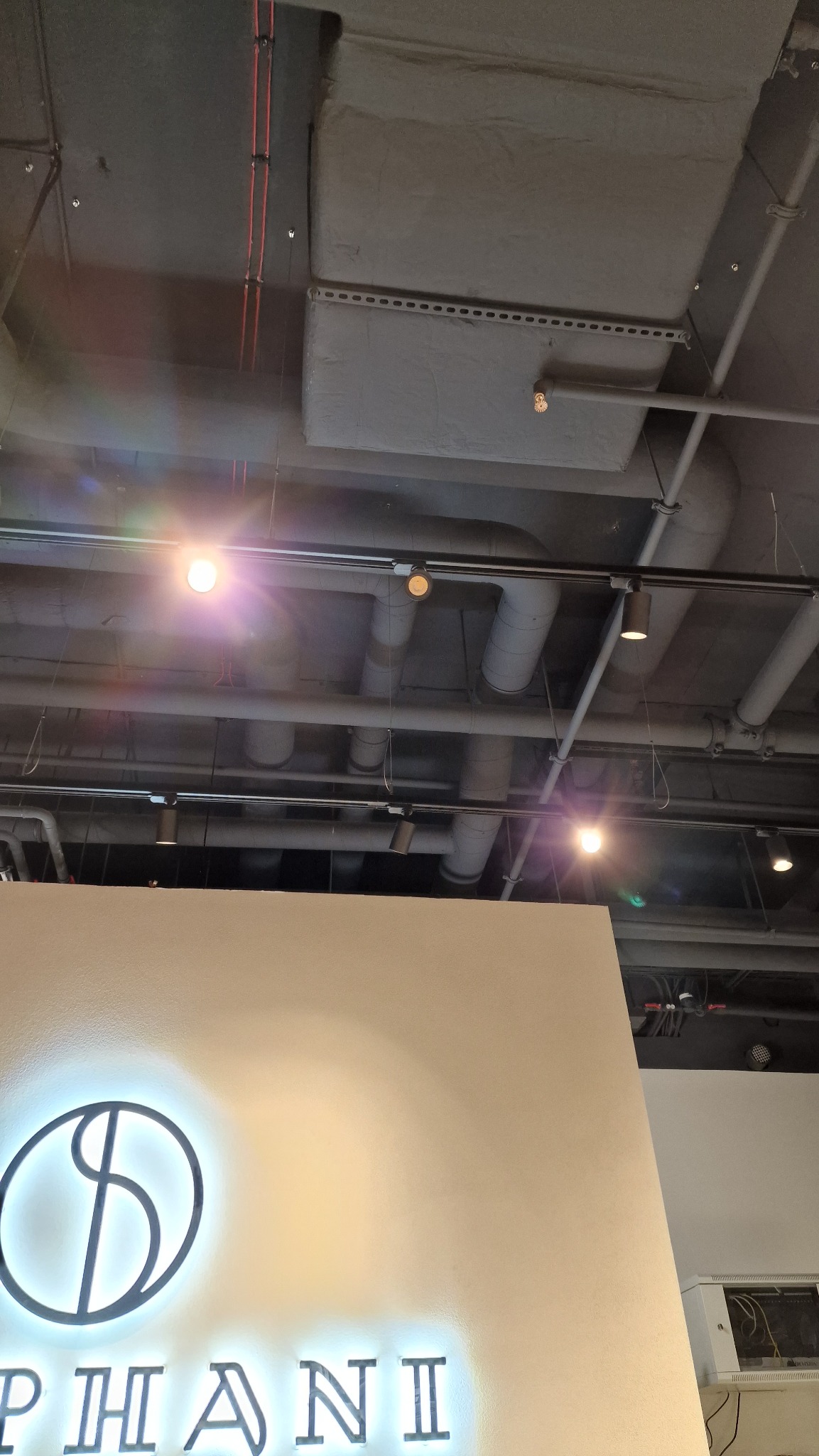 Szare rury wentylacyjne i oświetlenie sufitowe w biurze, widoczny fragment podświetlanego logo firmy na ścianie.