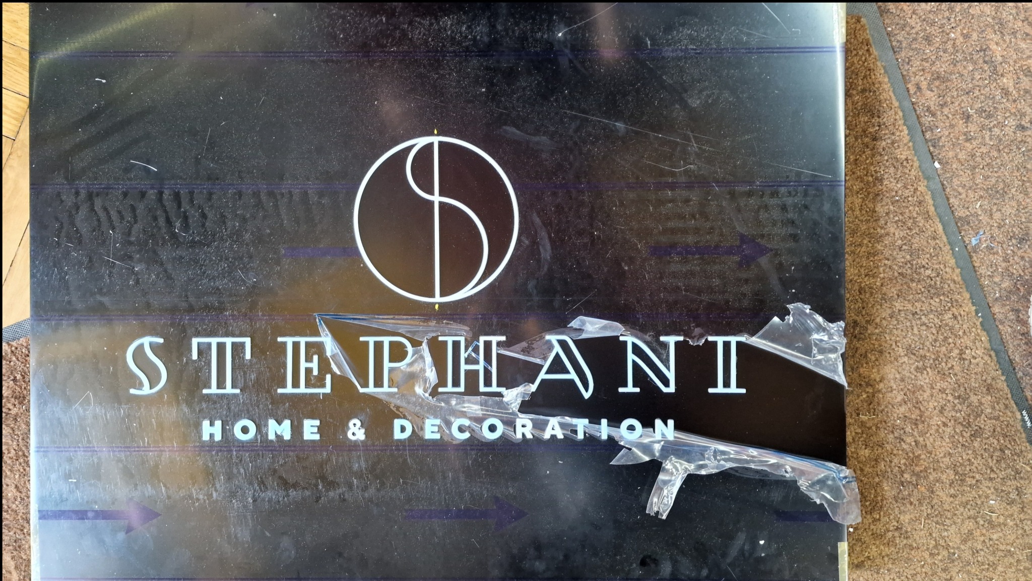 Błyszcząca czarna tablica z logo i napisem 'STEPHANI HOME & DECORATION' częściowo zakryta przez odrywającą się folię ochronną, leżąca na brązowej wykładzinie.