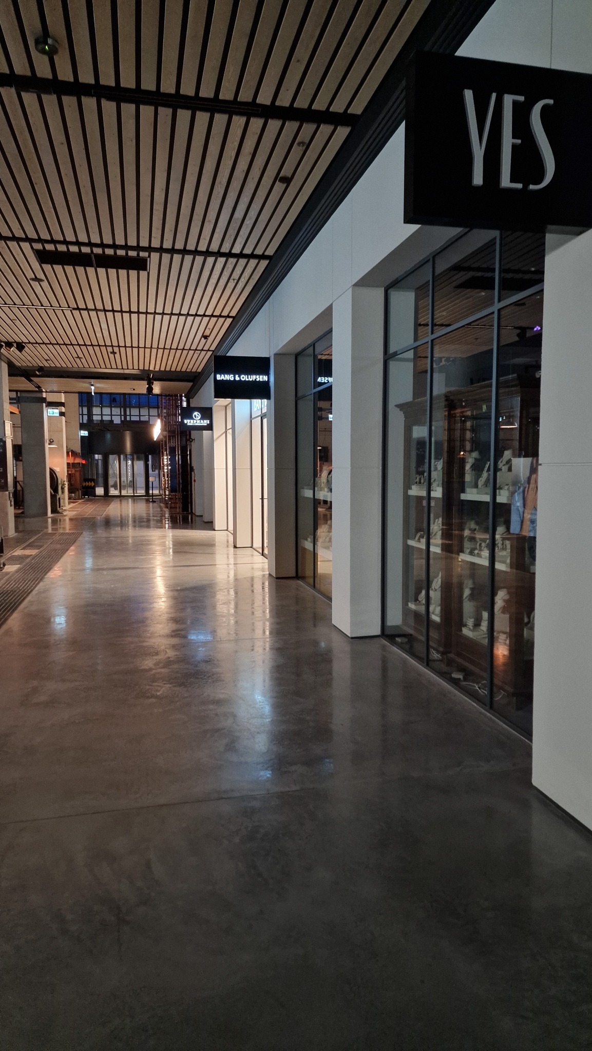 Polerowana betonowa posadzka w korytarzu biurowym z widocznymi sklepami odzieżowymi i elektronicznymi, podwieszany sufit z drewnianych listew.