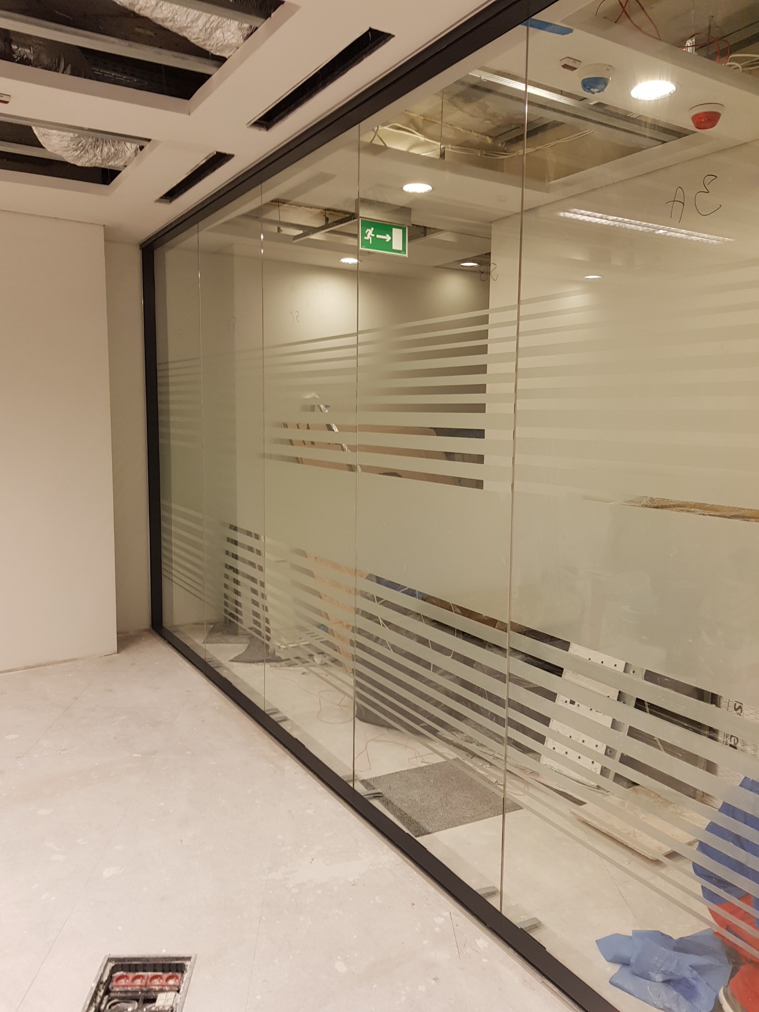 Szklana ściana działowa z poziomymi, matowymi pasami w biurze w trakcie remontu, widoczne niedokończone elementy sufitu i podłogi.