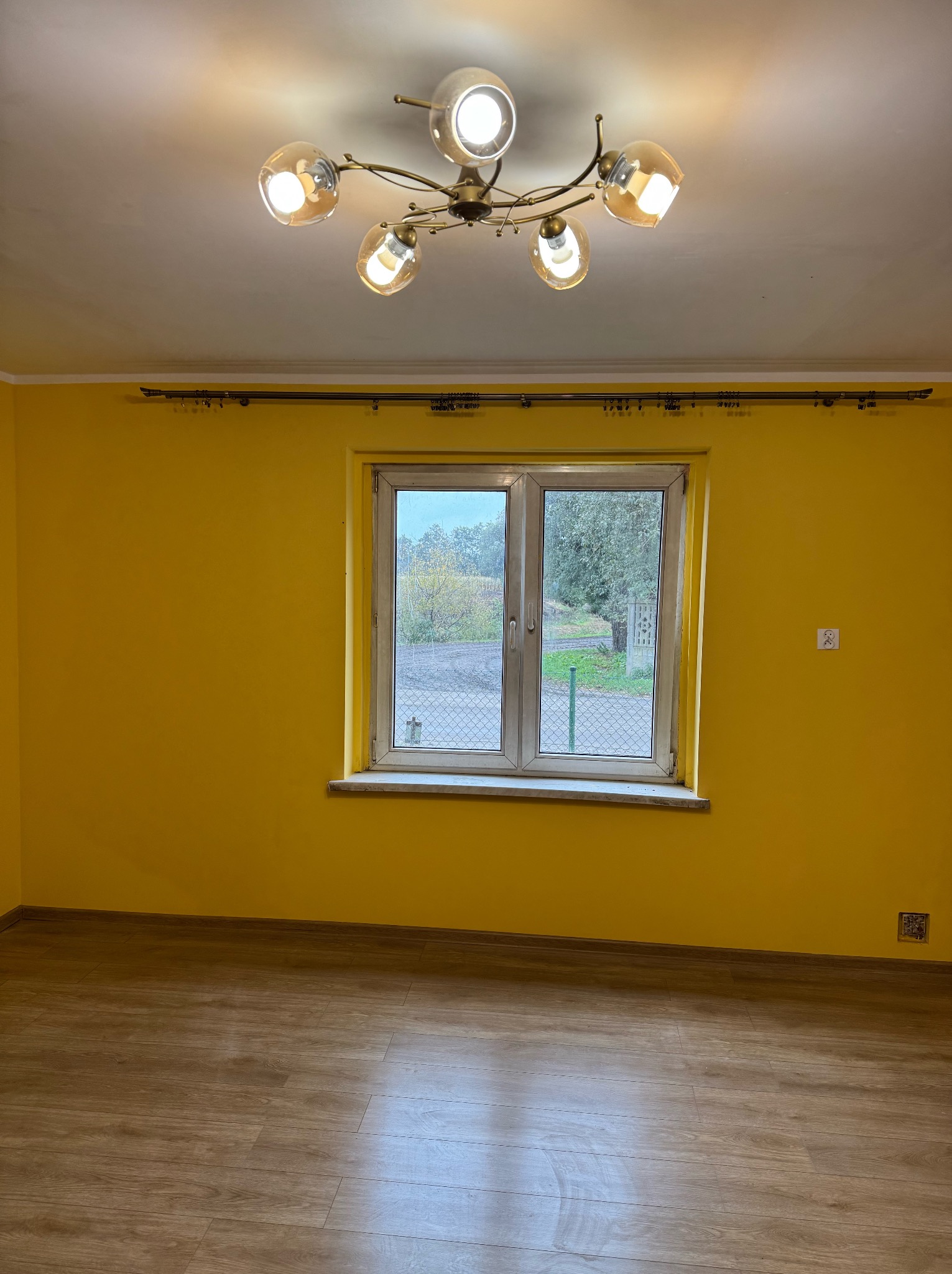 Pusty pokój z żółtymi ścianami, drewnianą podłogą i oknem z widokiem na zewnątrz, oświetlony przez lampę sufitową z sześcioma szklanymi kloszami.