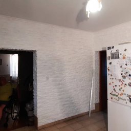 DUDA FINISH - Wykończona kuchnia z białymi ścianami imitującymi cegłę, szare szafki kuchenne, biała lodówka z magnesami i osobą na wózku inwalidzkim widoczną w przejściu do innego pomieszczenia.