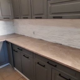 DUDA FINISH - Szare szafki kuchenne z czarnymi uchwytami, beżowy blat i ściana wyłożona dekoracyjną białą cegłą, widoczny środek do czyszczenia okien