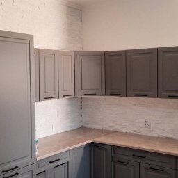 DUDA FINISH - Szare szafki kuchenne z czarnymi uchwytami, biała ściana z imitacją kamienia, lodówka z magnesami w tle.